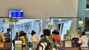 The image shows a group of people at an airport boarding gate, preparing to board a flight. Some of the individuals appear to be interacting with airport staff members, who are dressed in uniforms with official badges. Signs above the counters display "Boarding Economy" with instructions for passengers. In the background, a walkway to the aircraft is visible, along with a digital screen showing flight information. The scene captures a busy yet orderly boarding process at what appears to be an Indonesian airport.