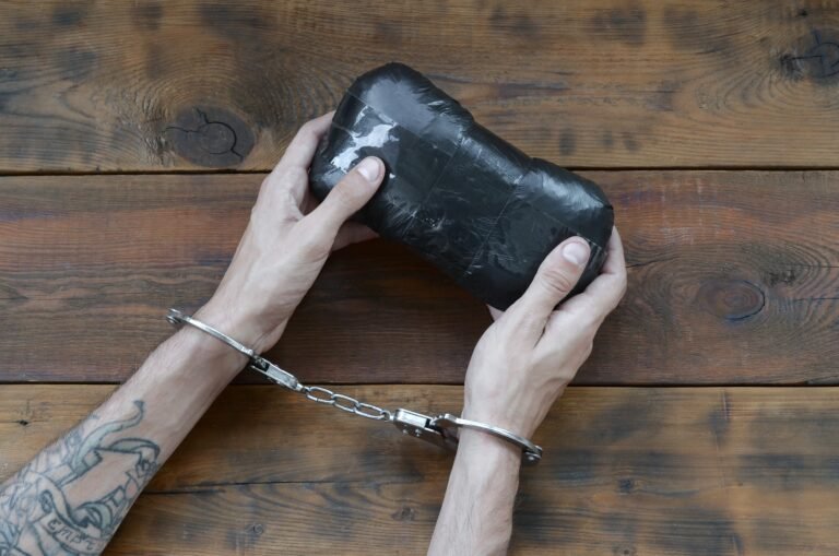 Arrested drug dealer hands in police handcuffs with big heroin drug package on dark wooden table background